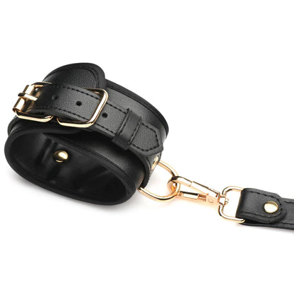 Nærbillede af en af de medfølgende manchetter til Strict Bondage Harness. Manchetten er lavet i sort vegansk PU-læder og har guldfarvede dele i nikkelfri metal. Lukkes som et bælte. Monteres på harness med medfølgende karabinremme. 